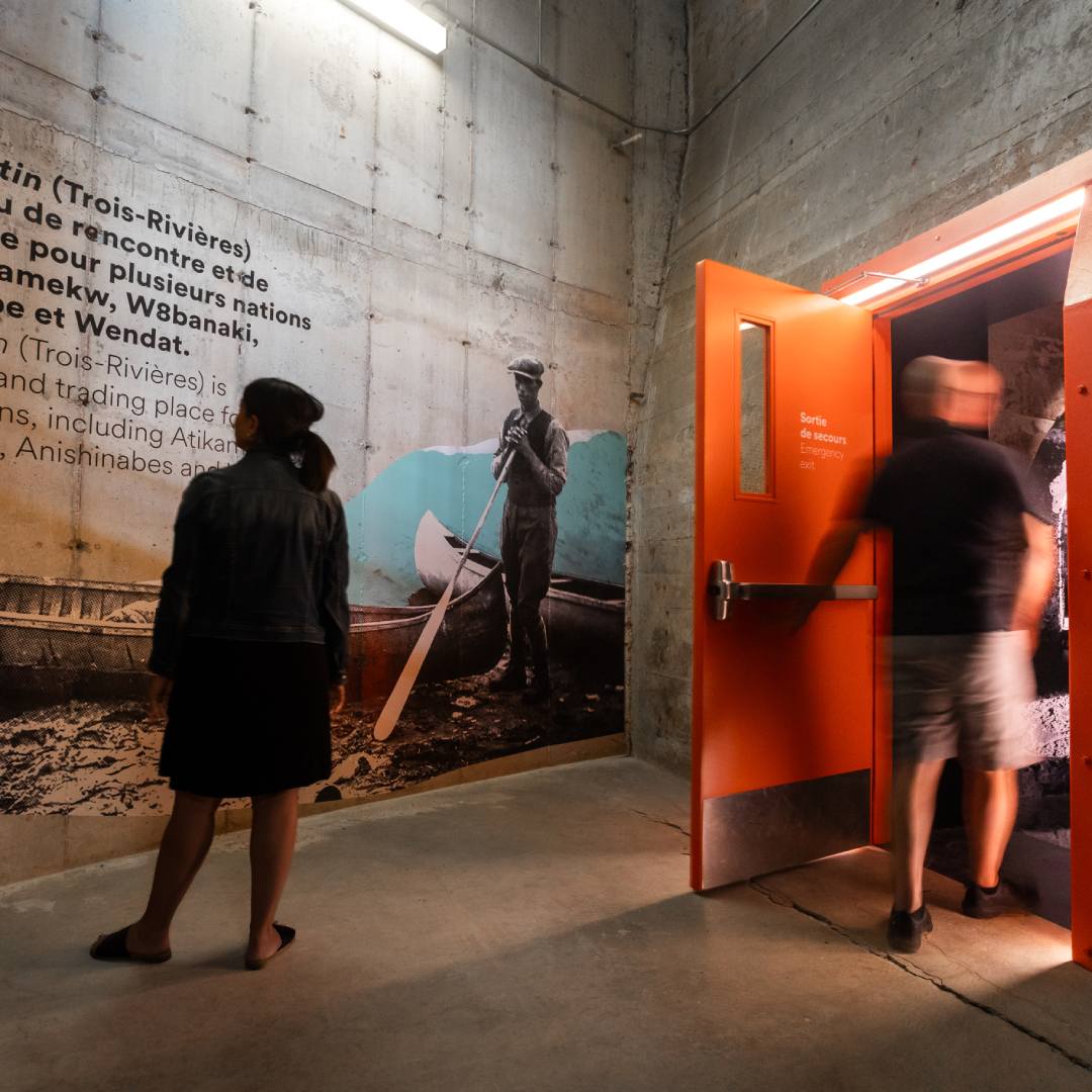 Une visiteuse observe une murale historique à l’intérieur du musée Boréalis, près d’une porte de sortie orange vif. On y voit un homme debout devant un canot.