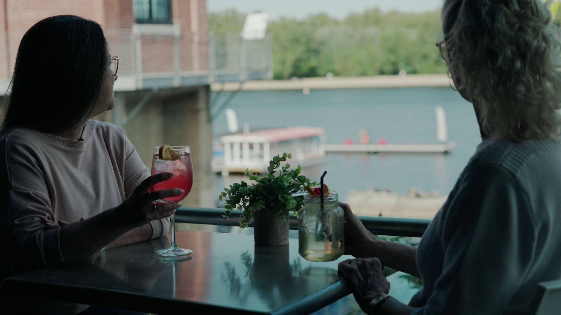 Au restaurent Le Castel, deux femmes discutent en sirotant des boissons colorées à une table près de la fenêtre, avec vue sur la rivière et la tour du musée Boréalis.