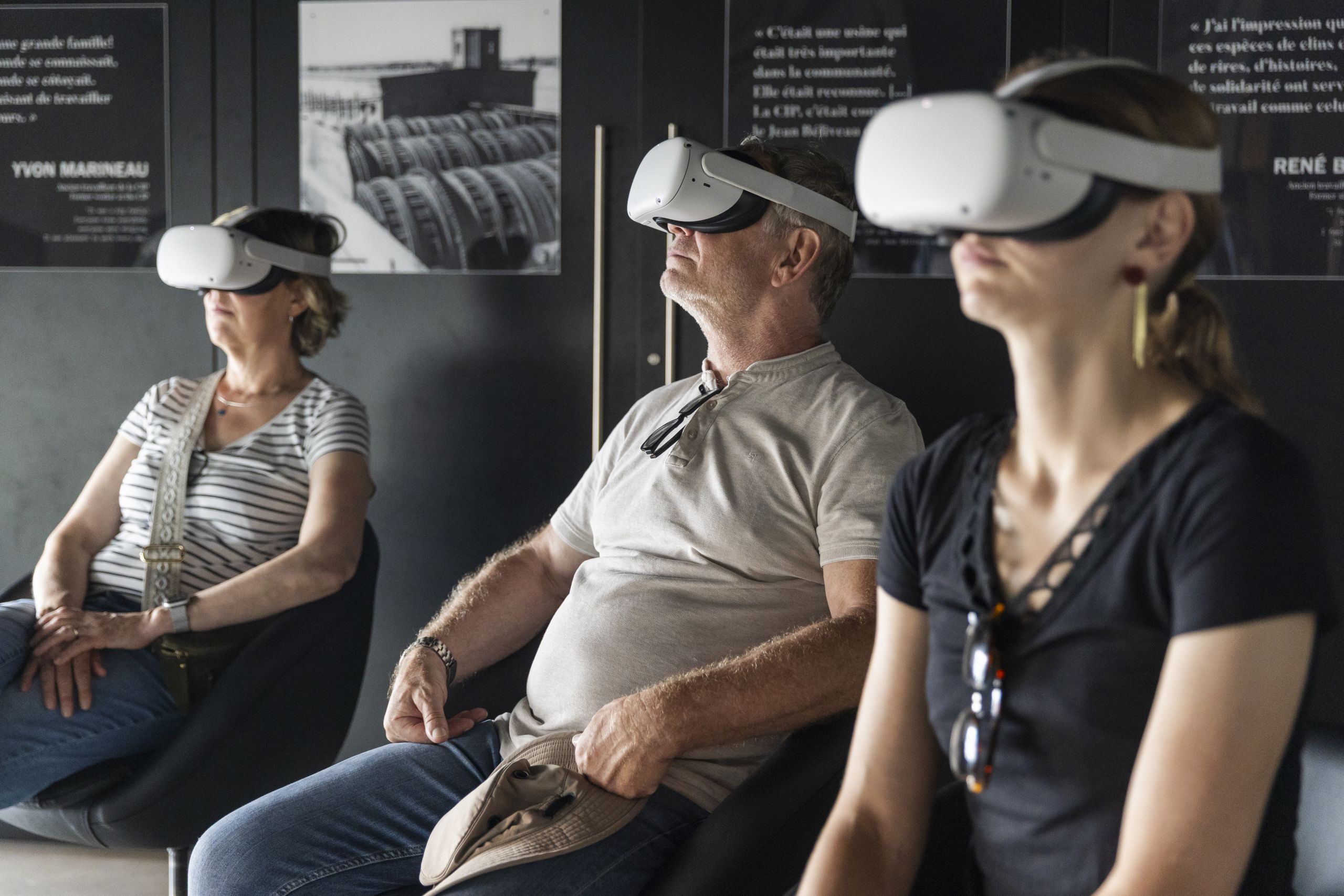 Trois personnes assises dans des fauteuils noirs portent des casques de réalité virtuelle. Leurs visages sont tournés vers le haut, concentrés, plongés dans l’expérience immersive Ma Belle St-Maurice. En arrière-plan, des photos et citations sont visibles sur un mur sombre.