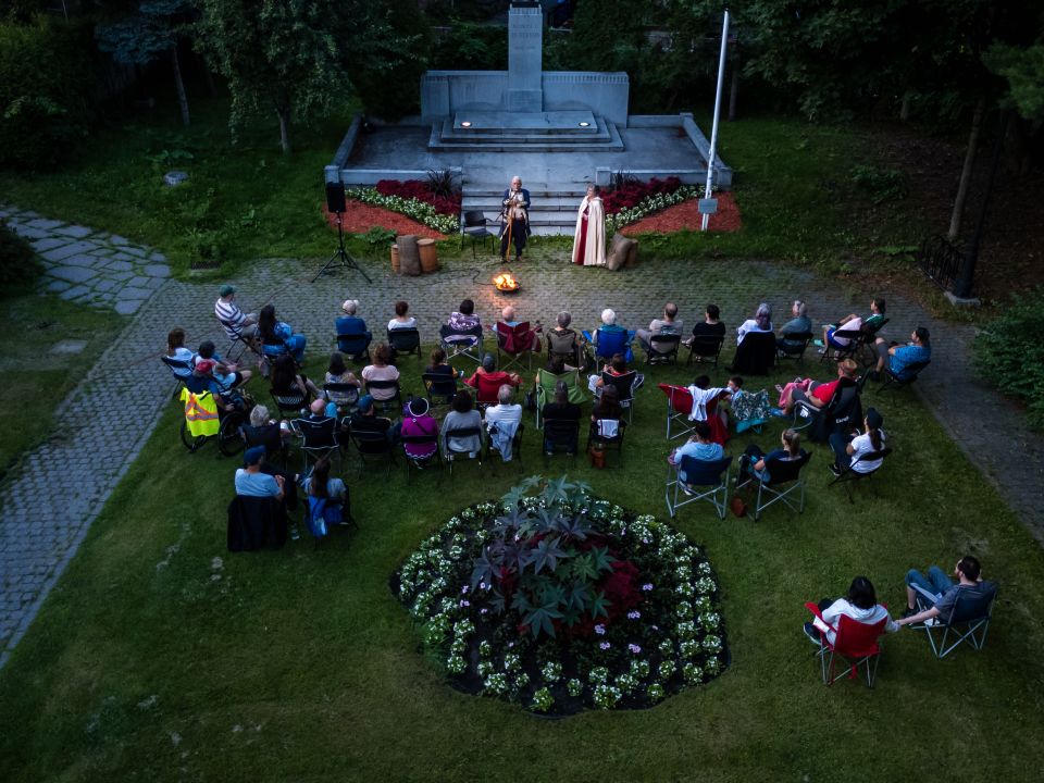 Une trentaine de personnes assises en demi-cercle sur des chaises de camping assistent à une soirée de contes autour du feu plein air, face à deux conteurs costumés. La scène se déroule au Manoir Boucher de Niverville.