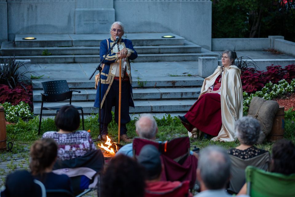 Un homme, debout avec une canne et vêtu d’un costume d’époque bleu, raconte une histoire devant un feu, face à un public assis. À ses côtés, une femme en cape crème et robe rouge est assise, attentive. En arrière-plan, le Manoir Boucher de Niverville encadrent la scène du conte.