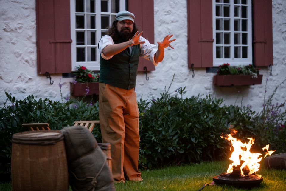 Le conteur Jean Philippe Marcotte, vêtu d’un pantalon orange, d’un gilet vert et d’une casquette, gesticule avec expressivité devant un feu de camp. Il se tient devant le Manoir Boucher de Niverville.